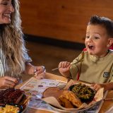 A woman is sitting with a little boy at a Sonny's BBQ dining table. She smiles looking at him as he prepares to take a bite of green beans from his plate.