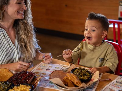 A woman is sitting with a little boy at a Sonny's BBQ dining table. She smiles looking at him as he prepares to take a bite of green beans from his plate.