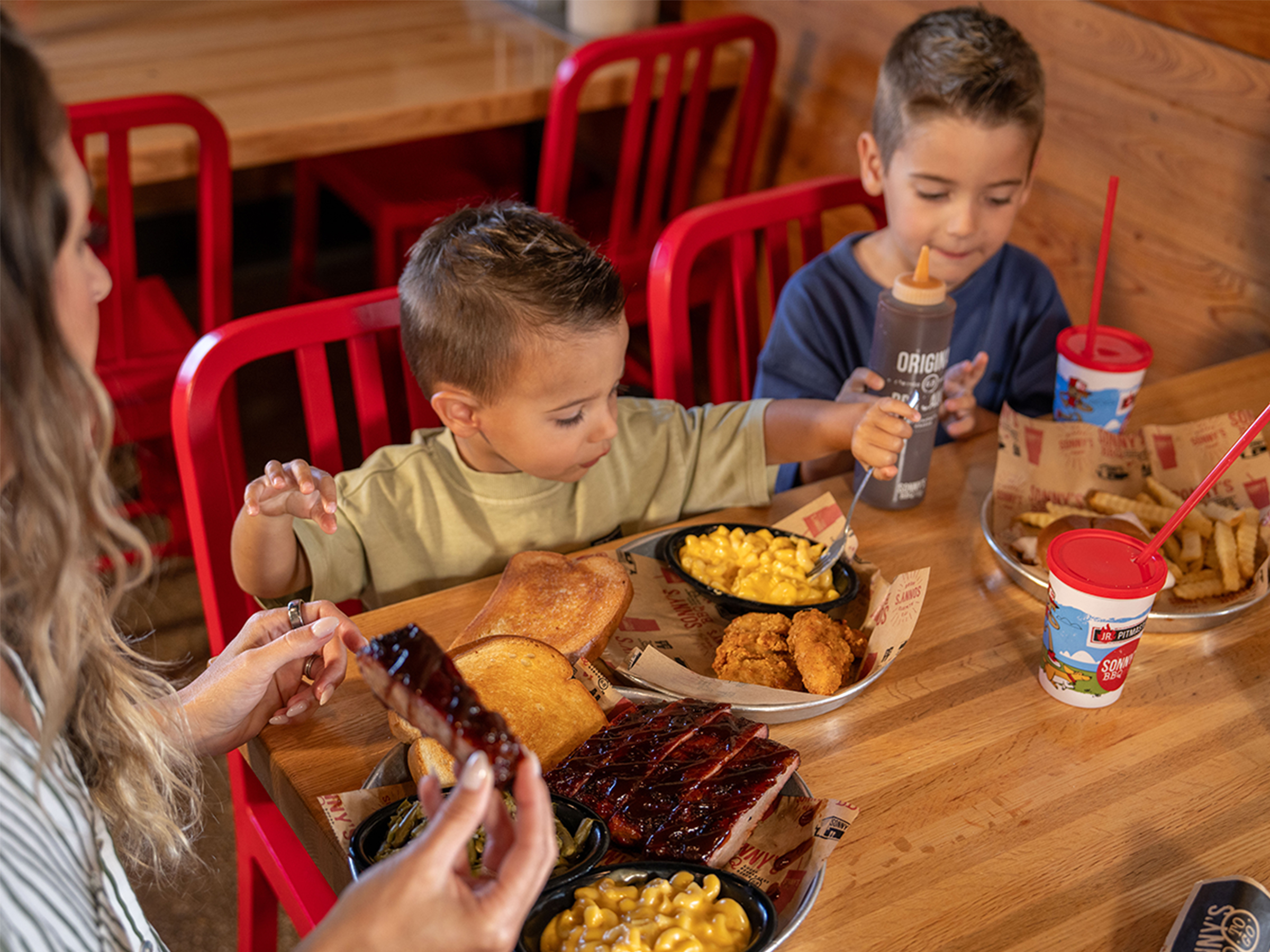 A woman and two little boys sit at a Sonny's BBQ dining table. Each one has a plate of food in front of them. The woman is holding a rib, and one of the boys is scooping mac and cheese.
