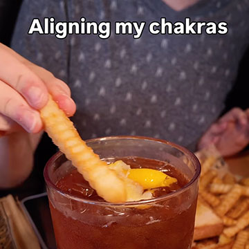 A person holding a french fry on the lip of a clear glass filled with sweet tea