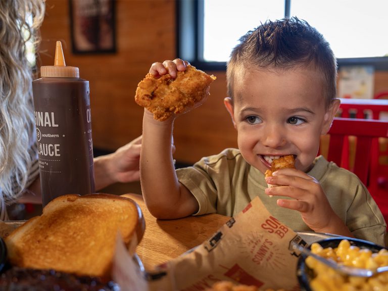 Little boy holds one chicken tender in one hand while biting into another with other hand. He sits at table filled with plates of BBQ and a bottle of Sonny's Original BBQ Sauce.