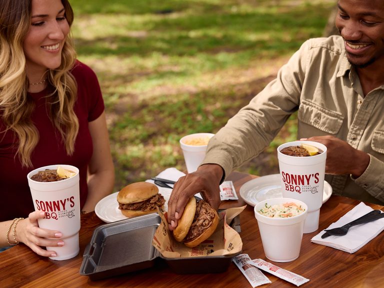 A man and woman sit at picnic table sharing a to go meal from Sonny's BBQ. The man reaches inside the to go box to grab a Pulled Pork Sandwich, while another sits on a plate nearby. The table is also filled with sides and drinks.