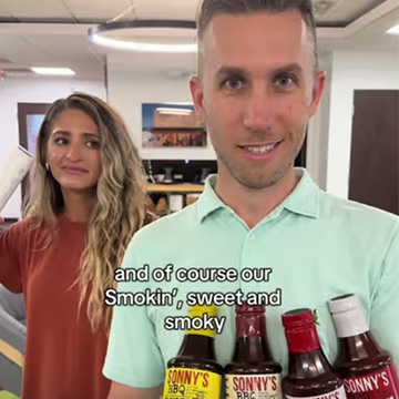 A man stands holding four bottles of BBQ sauce with a woman behind him.