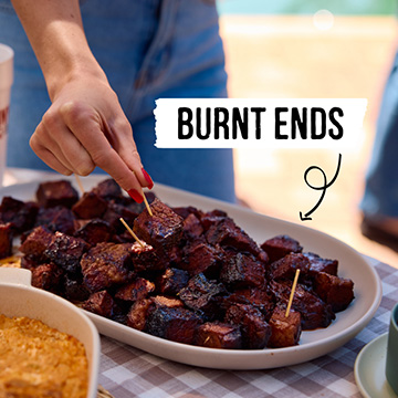 Woman grabs brisket burnt end from plate filled with them on checkered table.