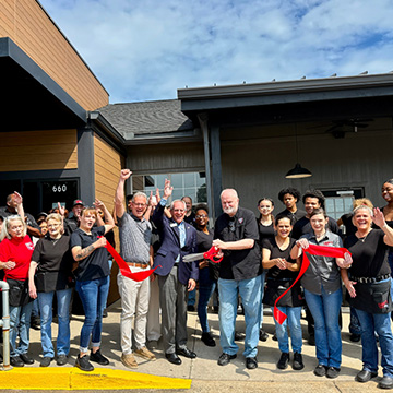 Group of people stand outside restaurant, cutting ribbon and celebrating