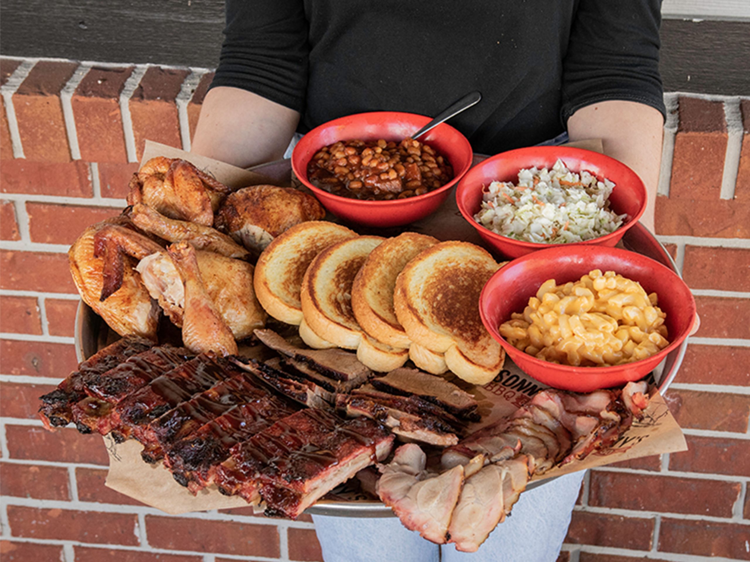 Person holding Sonny's Family Feast, a plate filled with Garlic Bread, Mac & Cheese, Coleslaw, BBQ Beans, BBQ Chicken, Sliced Pork, St. Louis Ribs, Brisket and Garlic Bread