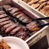 Tray of ribs sits next to tray of brisket. Tongs reach out to grab individual rib.