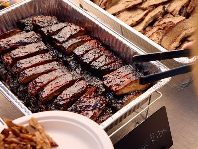 Tray of ribs sits next to tray of brisket. Tongs reach out to grab individual rib.