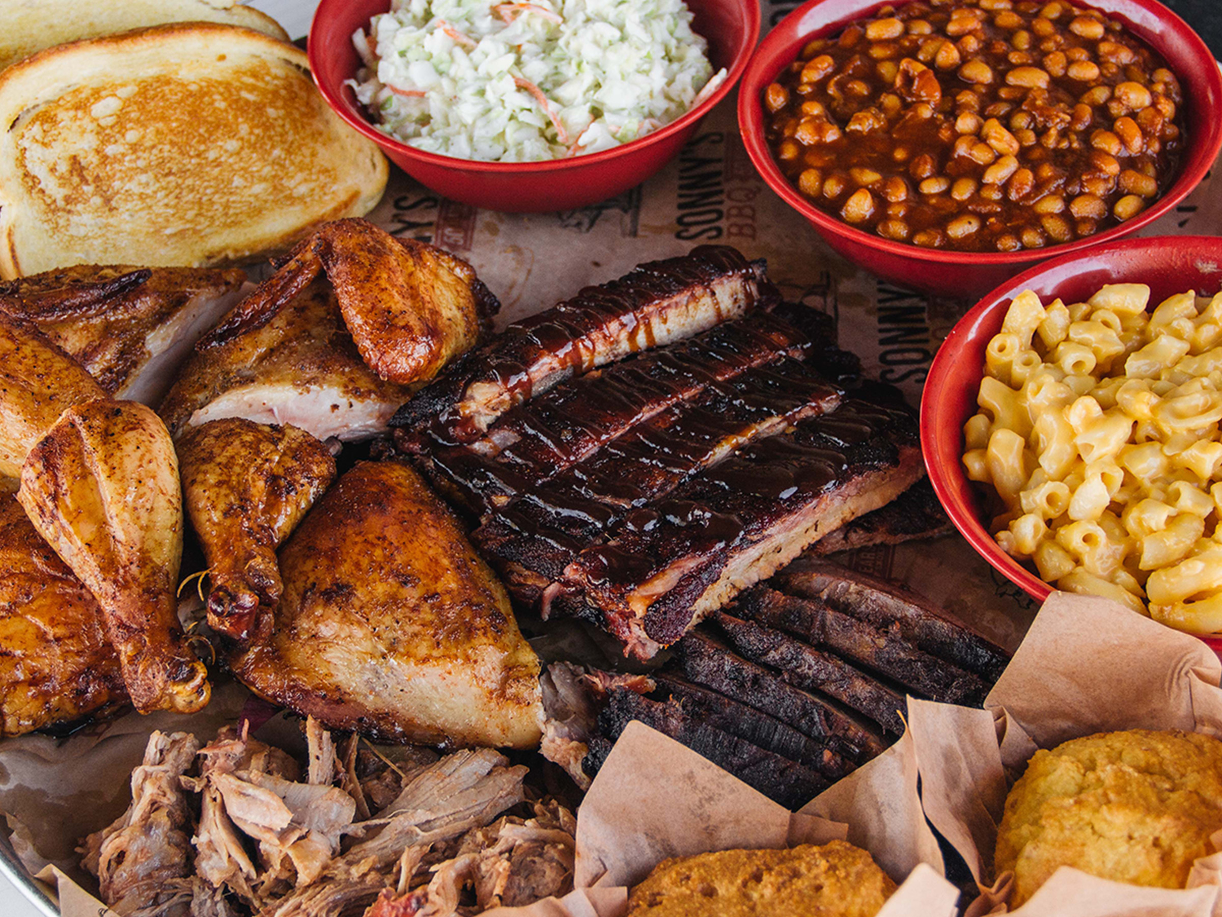 Close up shot of Sonny's Family Feast, including a platter filled with St. Louis Ribs, BBQ Chicken, Brisket, Pork, Garlic Bread, Cornbread, Coleslaw, BBQ Beans and Mac & Cheese.