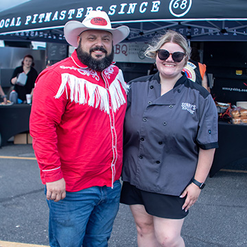 Man dressed in cowboy attire stands next to woman in Sonny's Catering shirt and shorts in front of Catering tent.
