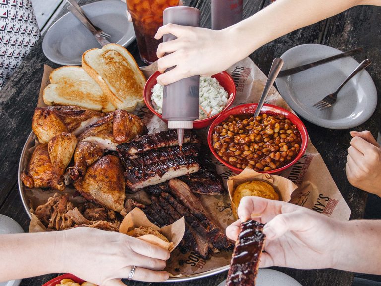 Multiple hands reaching over Sonny's Family Feast on table. Large Plate filled with BBQ beans, coleslaw, cornbread, garlic bread, chicken, ribs.