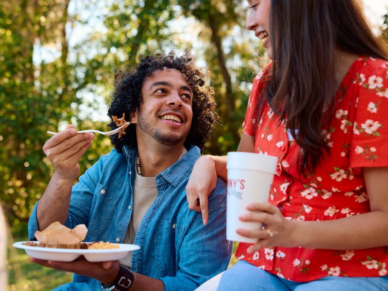 Smiling Man holds up fork of Pulled Pork to woman sitting next to him, while holding a plate full of BBQ in the other. Woman smiles at him while holding a Sonny's BBQ cup.