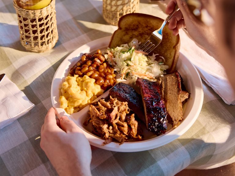 Plate of BBQ sits on checkered tablecloth. Plate contains Mac & Cheese, BBQ Beans, Coleslaw, Garlic Bread, Brisket, Ribs and Pulled Pork.