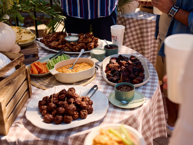 Multiple plates sit on decorated table with people surrounding it. Plates are filled with appetizers like BBQ Meatballs, Smoked Buffalo Chicken Dip and Smoked Wings.
