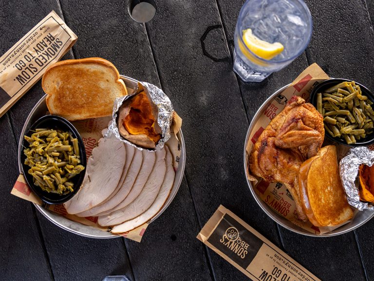 Two plates sit on a black picnic table, one filled with a Smoked Turkey Dinner and the other a Smoked Chicken Dinner. Two sets of silverware and glasses of water sit next to them. These are featured in the latest Sonny's BBQ nutrition blog.