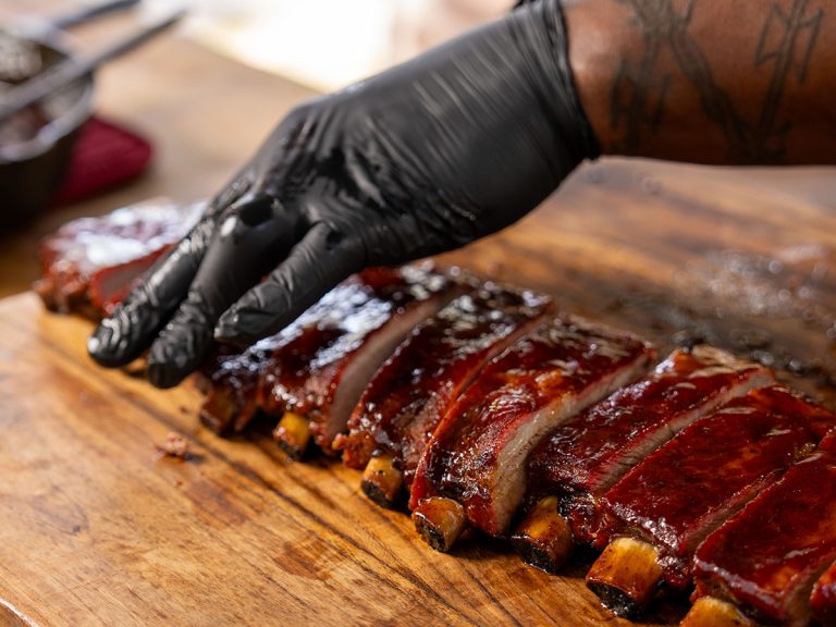 Gloved hand holds St Louis Ribs in place as knife slices them along cutting board