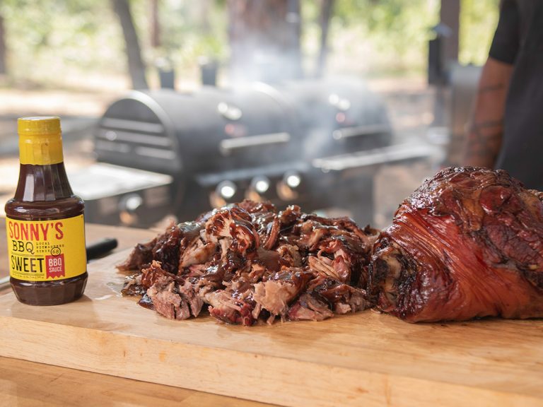 A bottle of Sonny's Sweet BBQ Sauce sits on a cutting board next to a pile of sauced Pulled Pork and a whole Pork Shoulder.