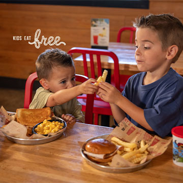 Two boys sit a table in front of two plates of food. One reaches to the other and tries to cheers with french fry. Text on picture reads 