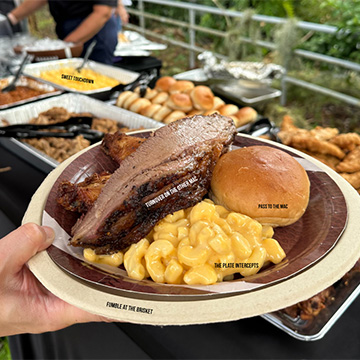 Plate full of Brisket, Mac & Cheese, Wings and a Roll is held up to camera. Small text dots the picture.