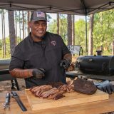 Man in Sonny's Pitmaster shirt points at Smoked Brisket that sits on table, which is sliced and sitting on a cutting board