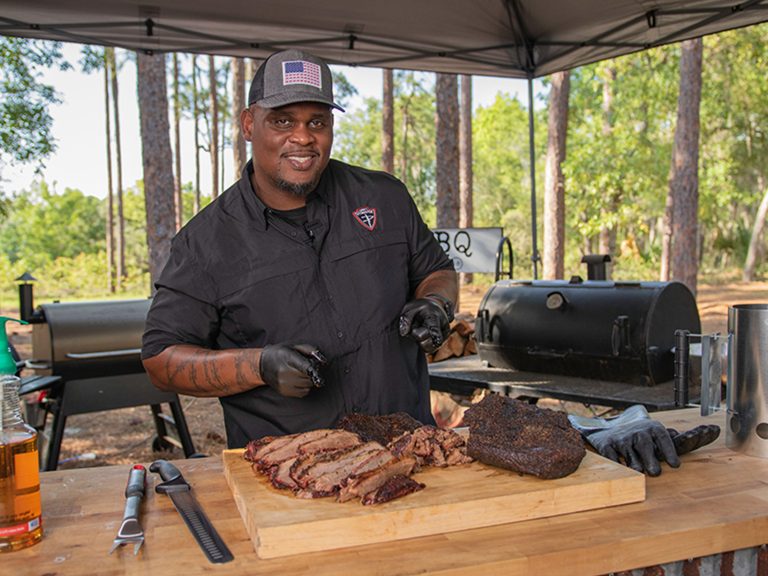 Man in Sonny's Pitmaster shirt points at Smoked Brisket that sits on table, which is sliced and sitting on a cutting board