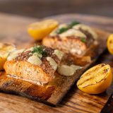 A tray of multiple filets of cedar plank salmon sits on a table along with grilled lemons.