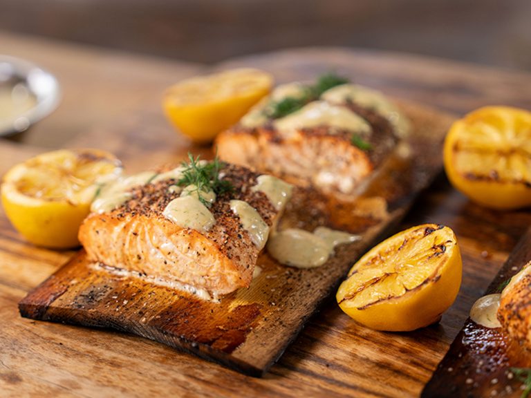A tray of multiple filets of cedar plank salmon sits on a table along with grilled lemons.