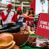 People enjoying BBQ outside football stadium, in front of them is a table lined with BBQ food and football decor. A book is superimposed on top which reads 