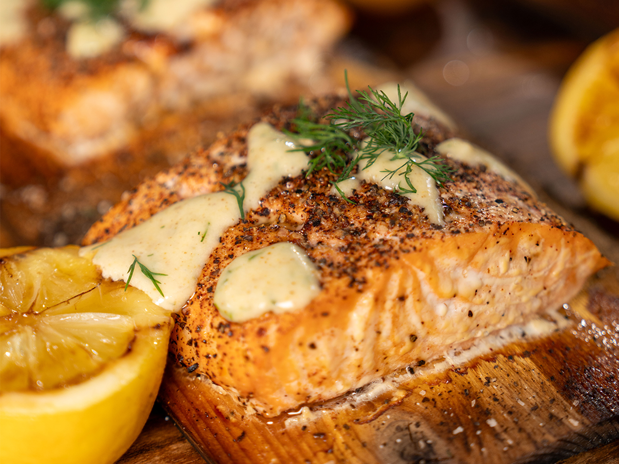 Cooked Cedar Plank Salmon sits on wood plank beside grilled lemons. It's topped with sauce and chopped dill.