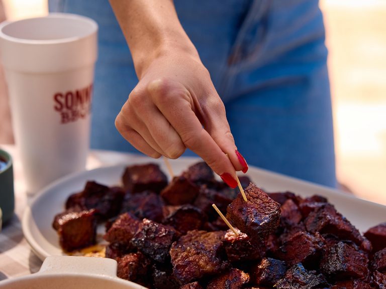 Woman's hand reaches onto plate to grab toothpick stuck into BBQ Burnt End.