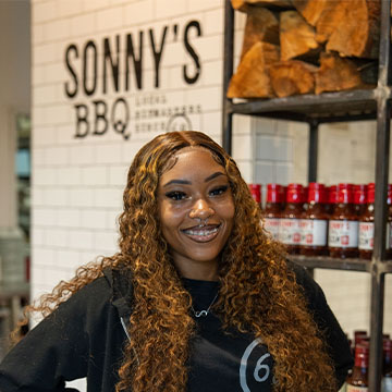 Young woman stands in front hallway of Sonny's BBQ, a shelf of sauces sits behind her.