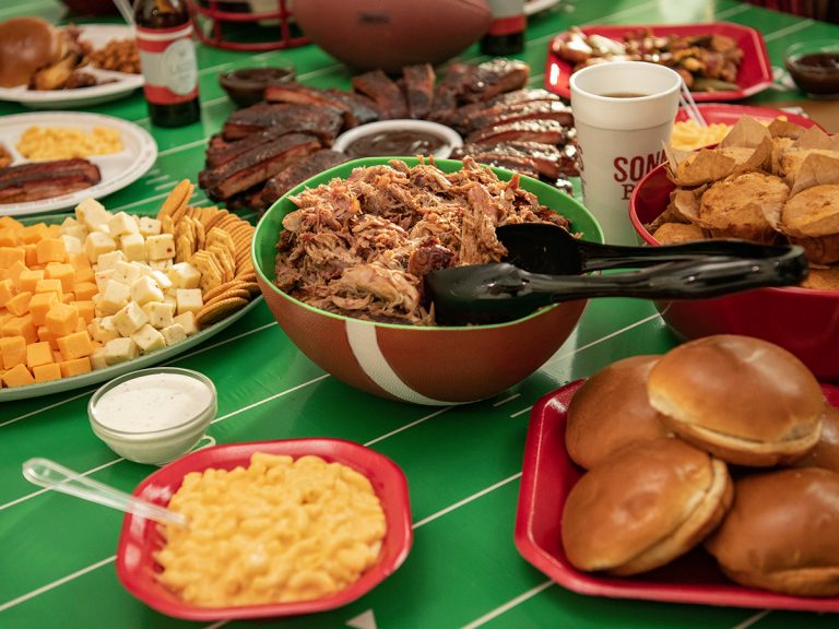 Tailgate BBQ Spread sits on table decorated with football decor. Food includes Pulled Pork, Cheese Cubes, Mac & Cheese, Cornbread and St. Louis Ribs.