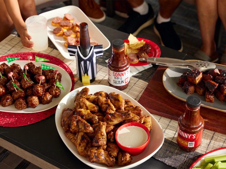Plates filled with BBQ Meatballs, Smoked Wings, Burnt Ends, and Pimento Cheese and Hot Link Crackers sit on coffee table. They're surrounded by bottles of sauce and football decor.