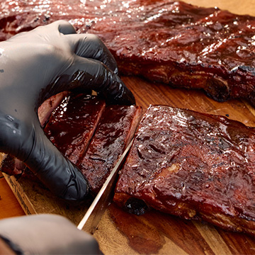 Two Slabs of St Louis Ribs sit on cutting board. One slab is being cut by a hand and knife in frame.