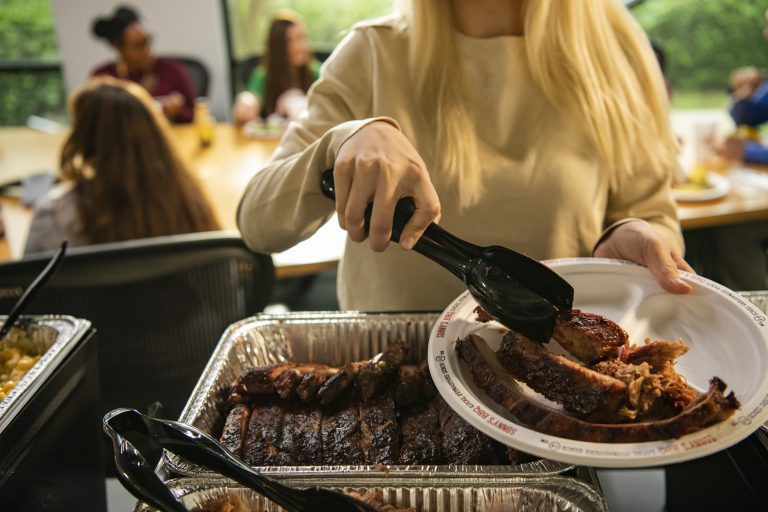 Woman in grabs rib from Sonny's Catering spread with tongs and places it on her plate. In the background is an office table.