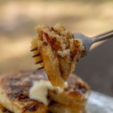 Fork holds pieces of multiple candied bacon pancakes up to camera, with stack of pancakes on plate sits in the background.