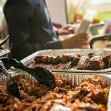In foreground, two tins of Sonny's Office Catering sit, one filled with Pulled Pork, the other with St. Louis Ribs. A man in the background is standing with a piece of paper in front of an office table filled with people.
