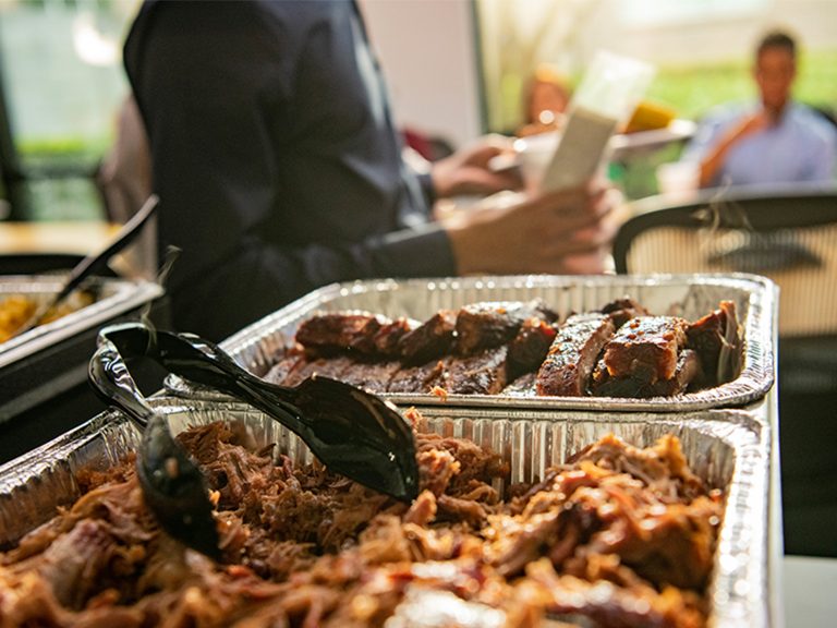 In foreground, two tins of Sonny's Office Catering sit, one filled with Pulled Pork, the other with St. Louis Ribs. A man in the background is standing with a piece of paper in front of an office table filled with people.
