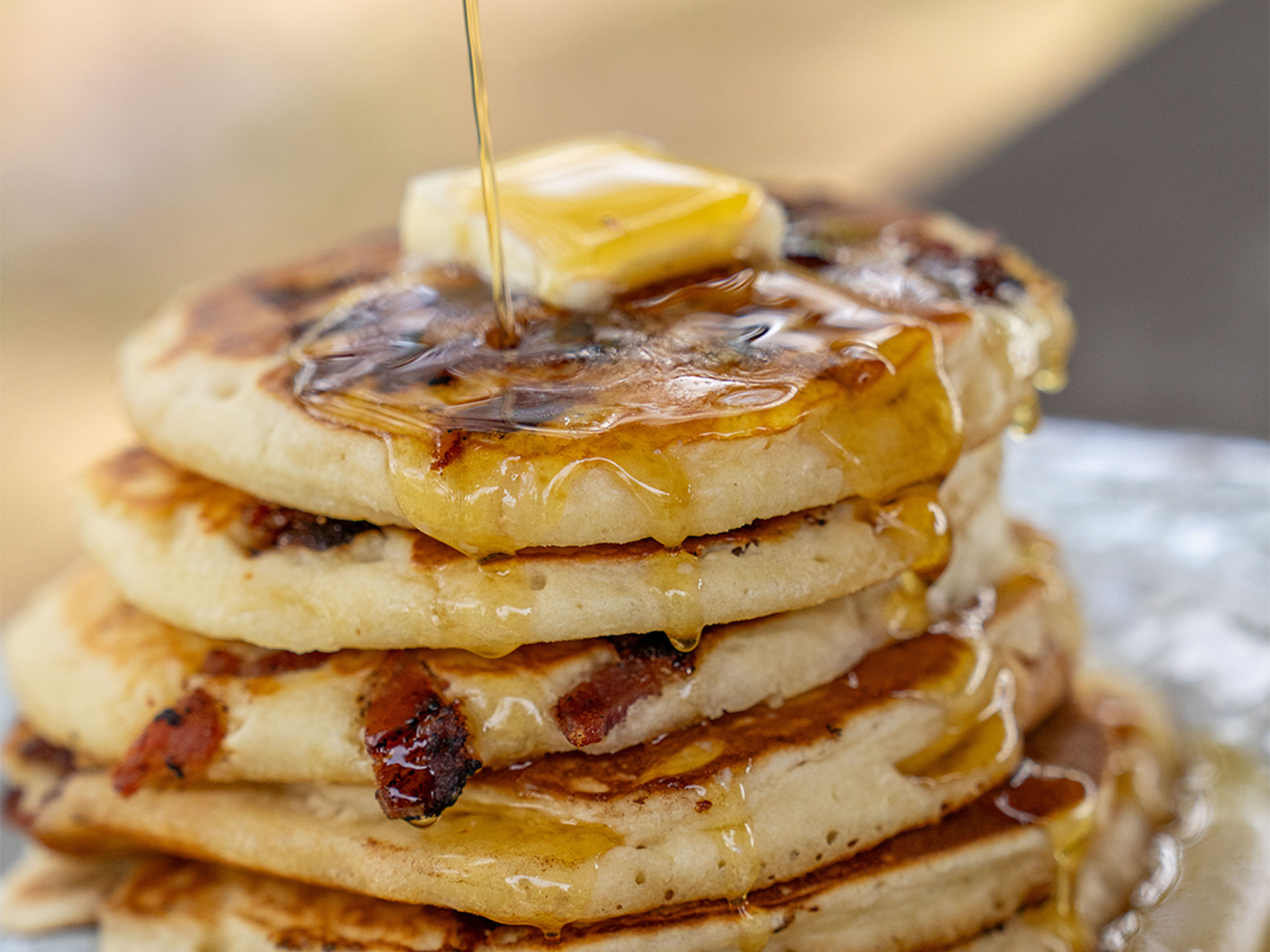 Stack of candied bacon pancakes topped with butter and dripping with syrup on a plate.
