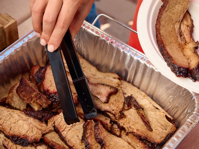 Hand holding tongs reaches in to grab slices of brisket from dish.