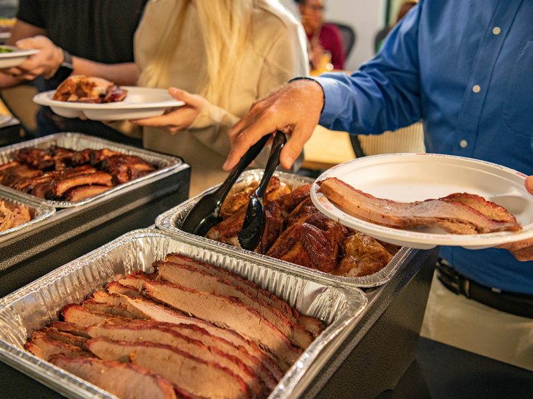 Man stands in front of office bbq catering setup, holding tongs and his plate, while grabbing a piece of chicken from a catering pan