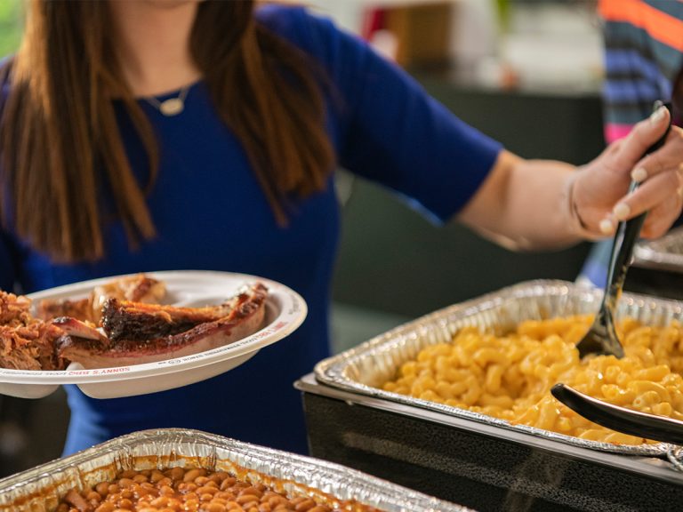 With one hand, woman in office scoops mac and cheese from pan with a spoon, while holding a plate with ribs and pulled pork on it in her other hand.