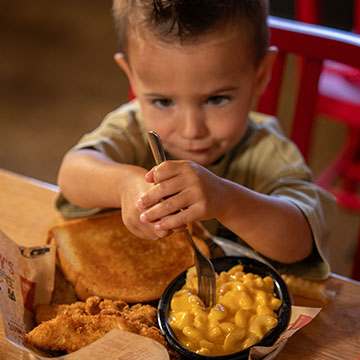 Little boy holds fork with two hands and tries to scoop mac & cheese off plate in Sonny's restaurant.