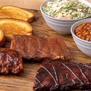 Three slabs of ribs sit on a cutting board beside Garlic Bread and two bowls of Coleslaw and BBQ Beans.