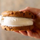 Hand holds cookie ice cream sandwich out in front of tan background.