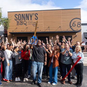 Man holds ceremonial scissors high in air and surrounded by cheering crowd at restaurant ribbon-cutting