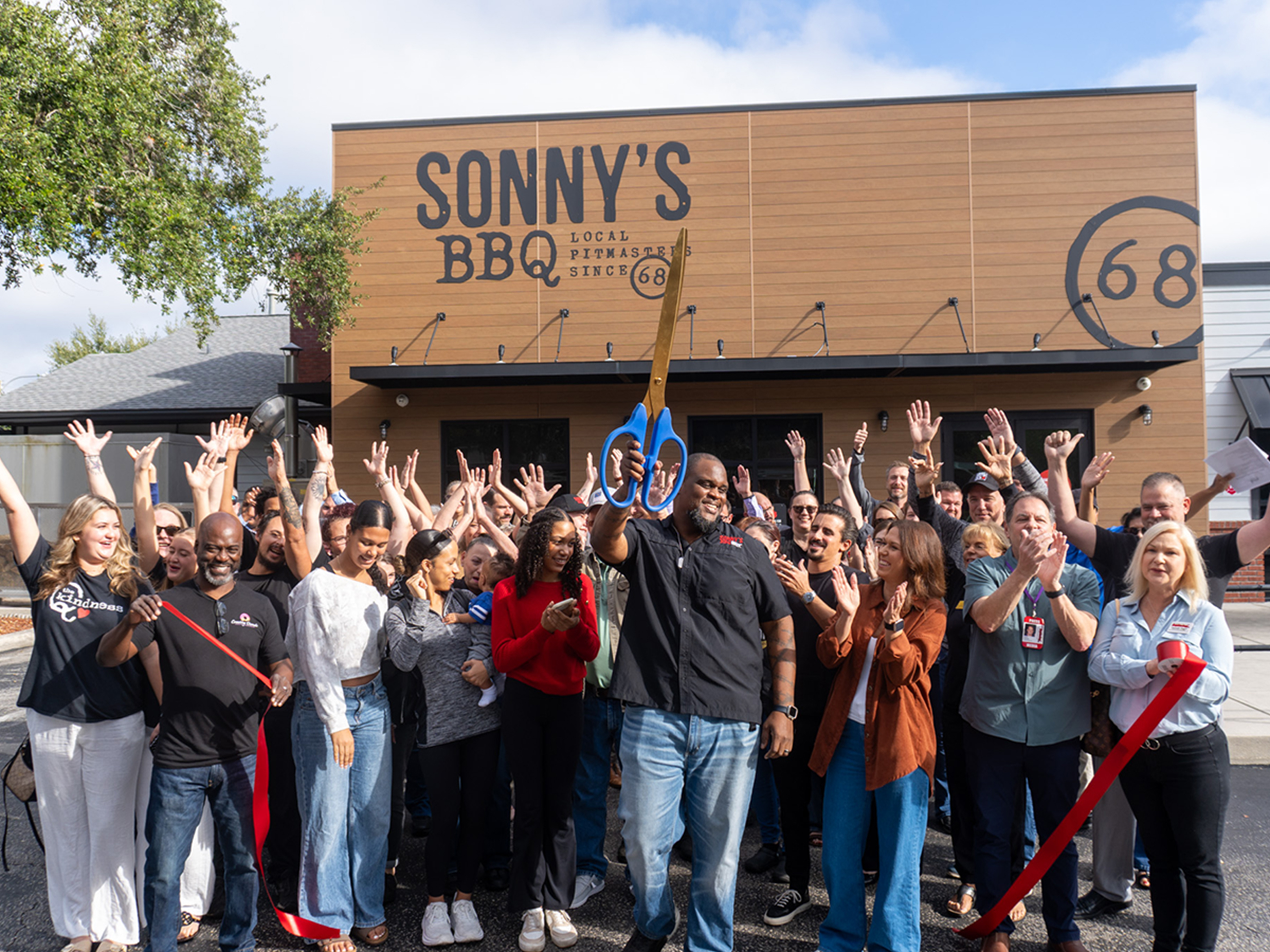 Crowd cheers as man holds up scissors at ribbon-cutting ceremony outside Sonny's