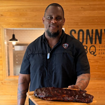 Pitmaster smiles at camera carrying cutting board with racks of cooked ribs