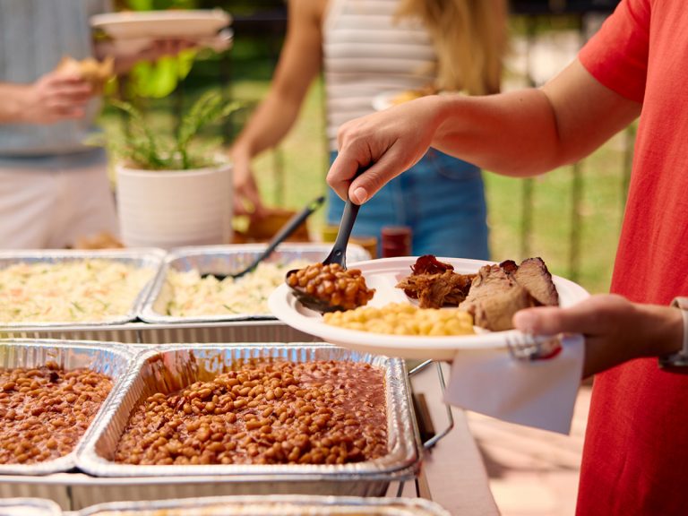 Woman grabs spoonful of BBQ Beans from catering spread for her plate.