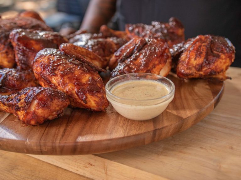 Multiple pieces of Smoked Chicken sit on cutting board alongside cup of dipping sauce